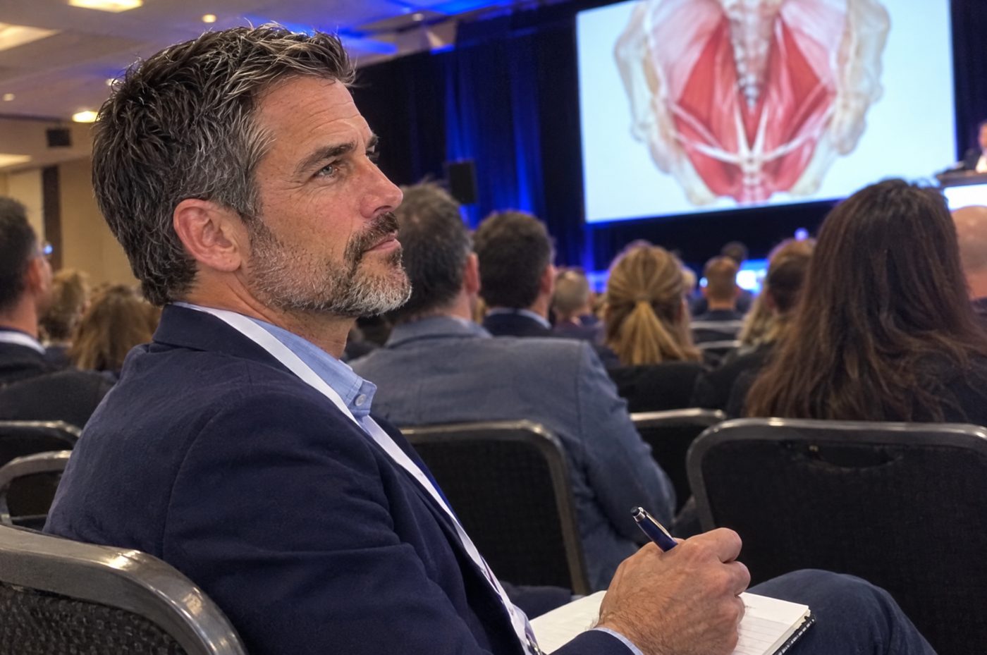 Dr. Mercer in the audience at a medical conference, an anatomy slide on screen