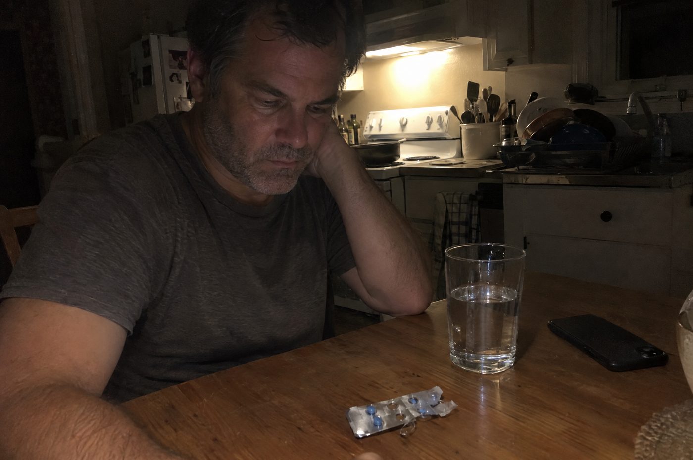 A man staring at a blister pack of blue pills at his kitchen table at night